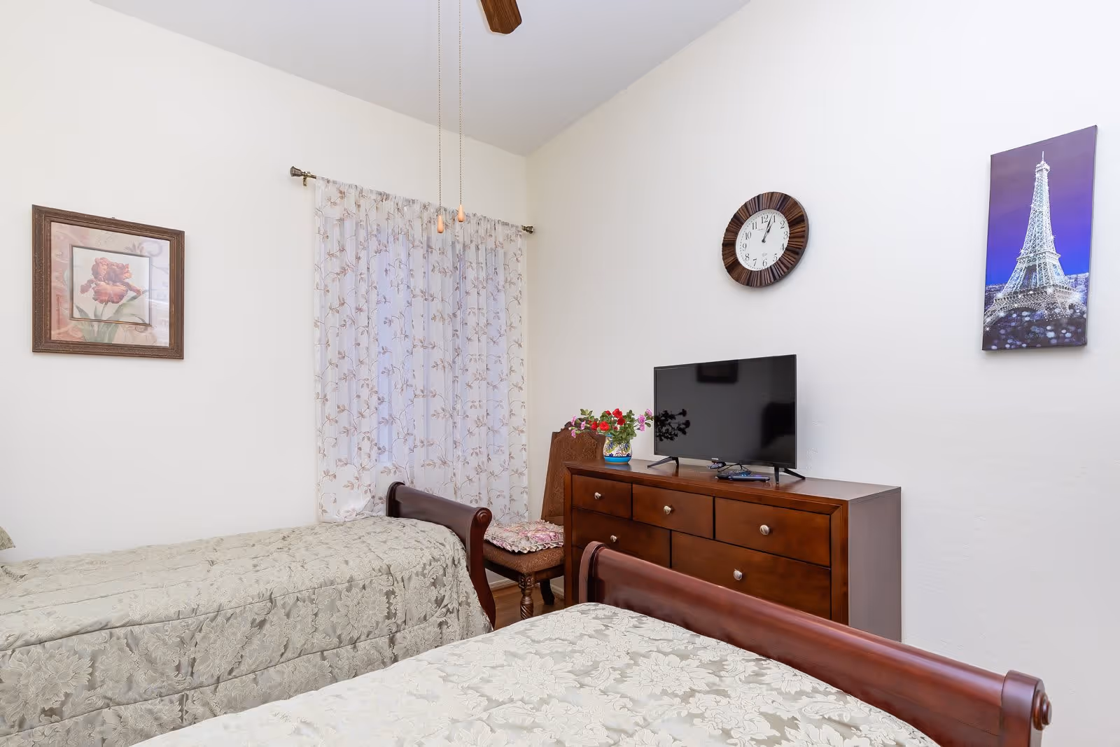 A bedroom with two twin beds covered in beige floral bedspreads. Between the beds is a wooden dresser with a flat-screen TV on top. A wooden chair with a floral cushion is placed next to the dresser. The walls are white, decorated with a framed flower picture, a round wall clock, and a picture of the Eiffel Tower. A window with sheer floral curtains is visible behind the chair.
