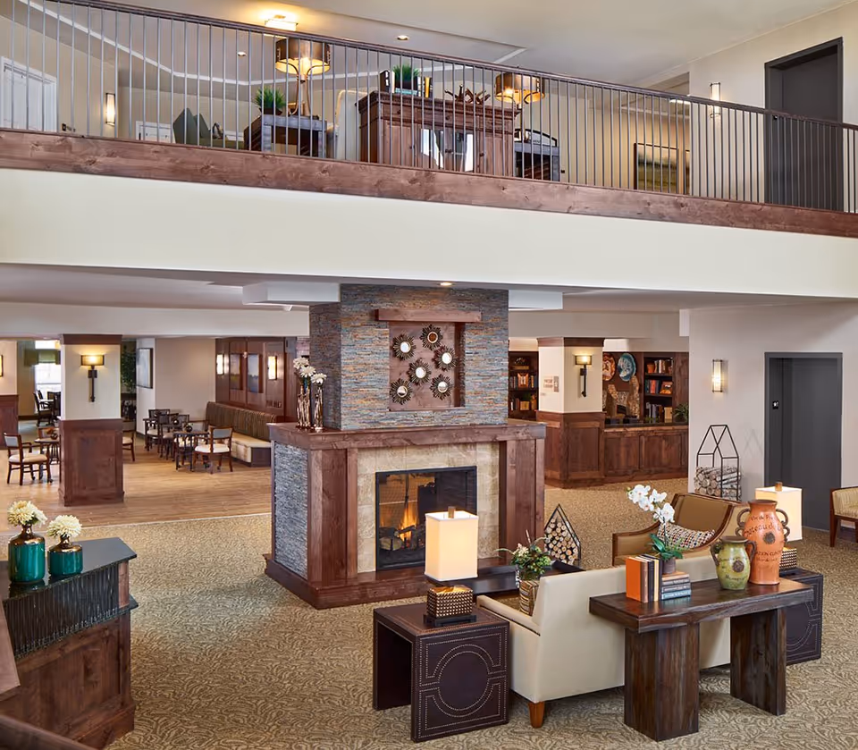 Bright two-level communal living area with seating, a central stone fireplace, decorative accents and an upper balcony.