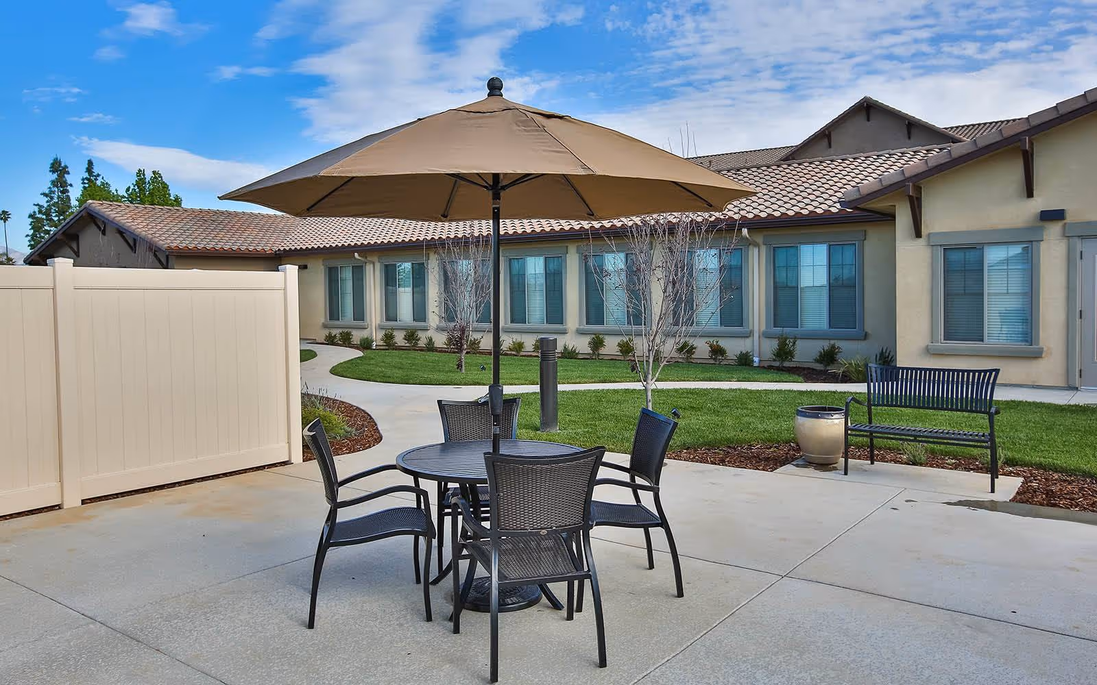 Courtyard patio with a round table, four chairs and umbrella in front of a single-story senior living building.