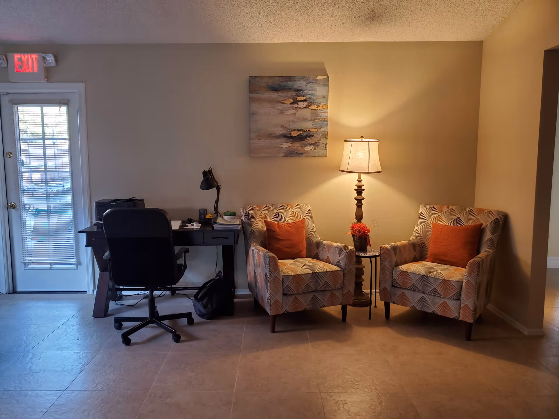 A cozy interior corner featuring two patterned armchairs with orange cushions separated by a small round table holding a lamp and a small flower pot. To the left, there is a black office chair in front of a dark wooden desk with a desk lamp, printer, and some office supplies. A door with window blinds and an illuminated exit sign is visible on the far left.