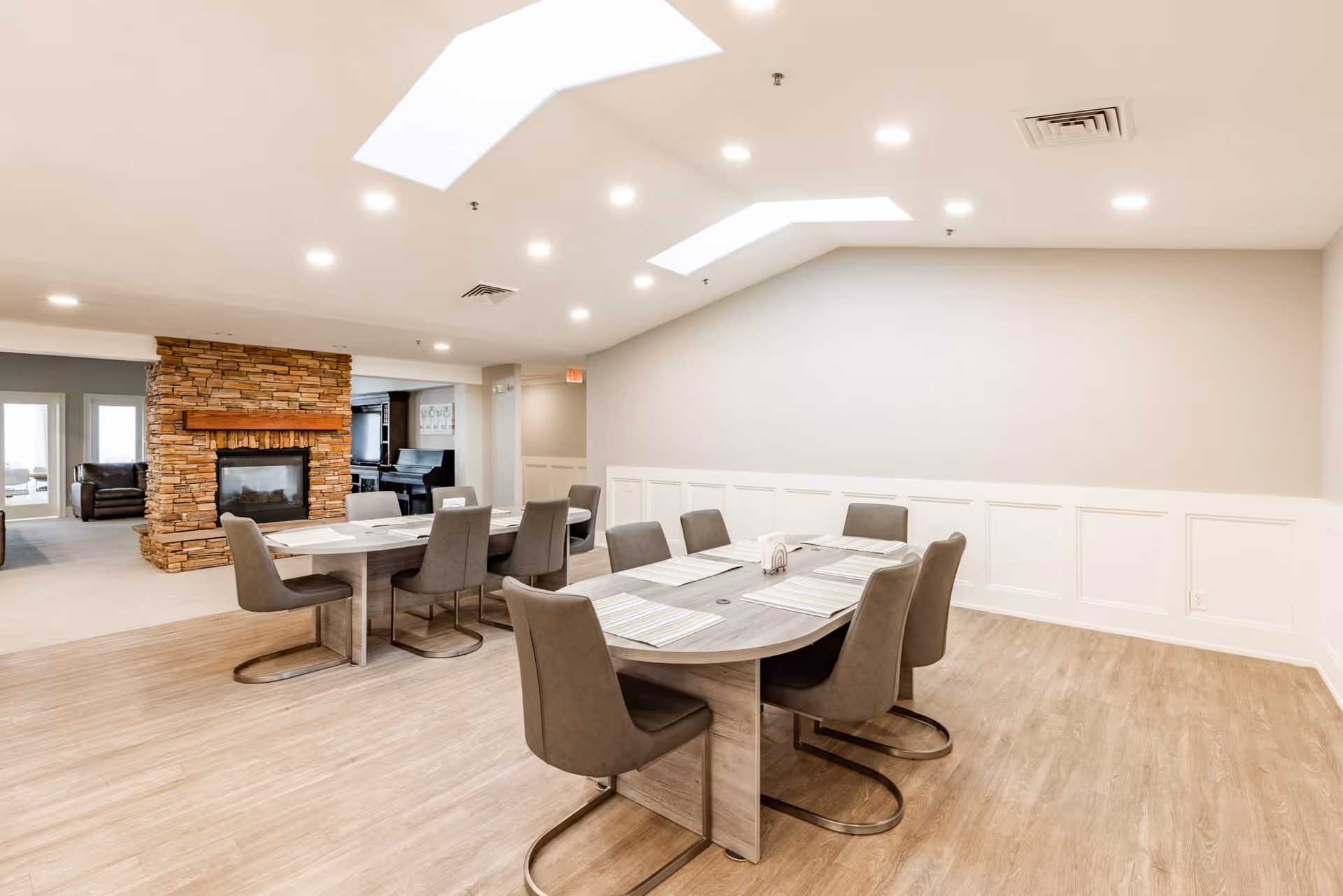 A bright and spacious dining area with a large wooden table surrounded by eight gray cushioned chairs. The room features light wood flooring, white paneled walls, and two skylights in the ceiling providing natural light. In the background, there is a stone fireplace and a piano in an adjacent room.