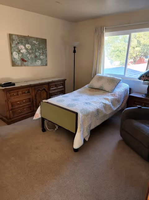 A small bedroom with a single bed covered with a white quilt with floral patterns. There is a wooden dresser with multiple drawers against the wall, a tall floor lamp in the corner, a window with cream curtains letting in natural light, and a cushioned armchair. A floral painting hangs on the wall above the dresser.
