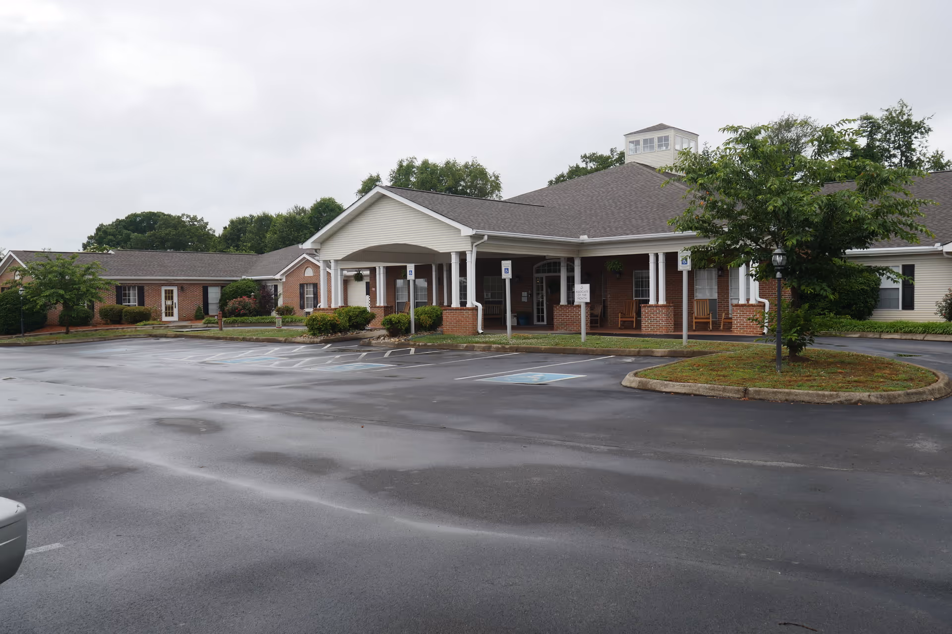 Front exterior of a single-story senior living building with a covered entrance, porch seating, and an empty wet parking lot.