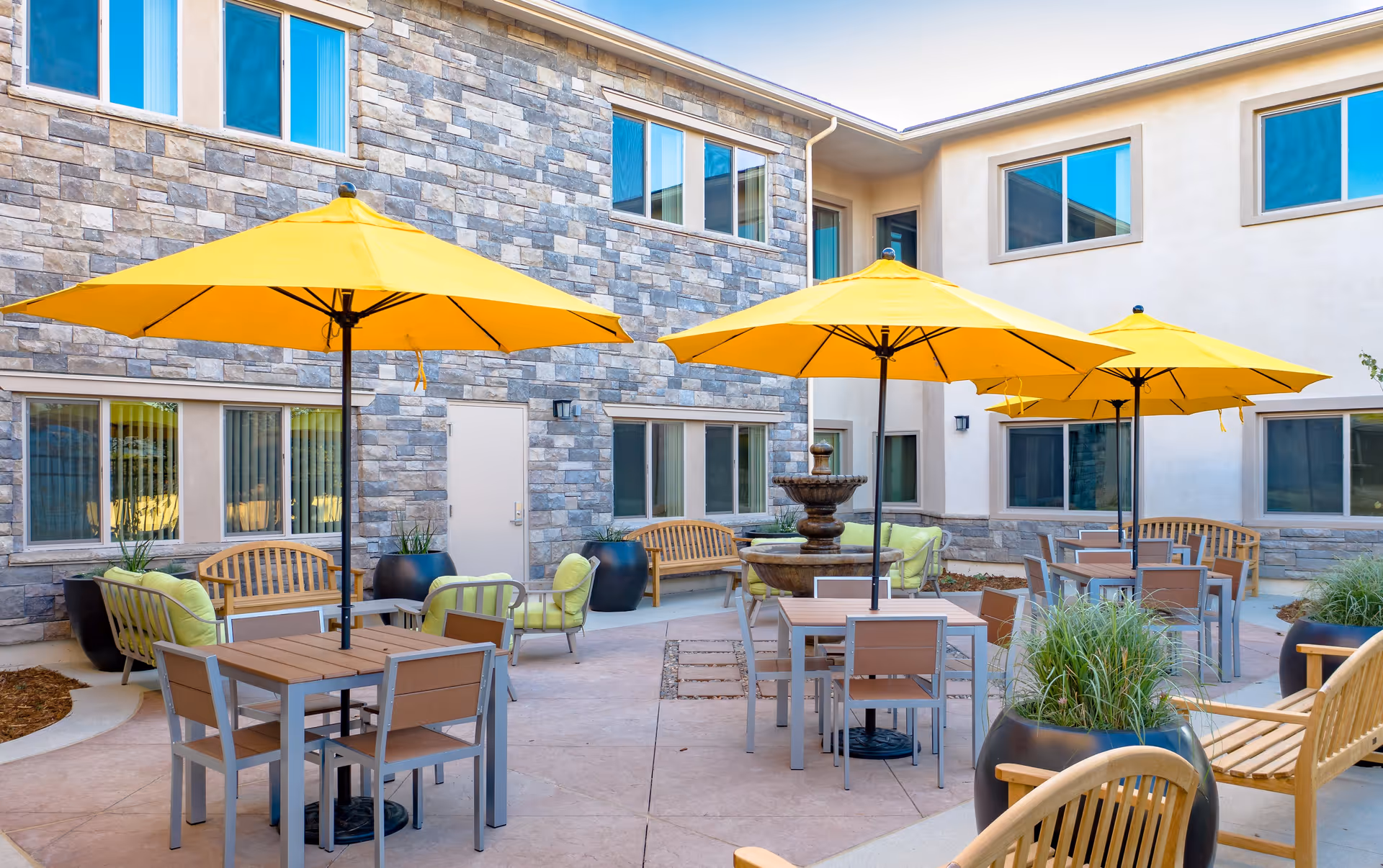 Outdoor courtyard area at Vista At Simi Valley featuring several tables with yellow umbrellas, wooden benches with green cushions, large potted plants, and a central stone water fountain, surrounded by a two-story building with stone and stucco exterior walls and multiple windows.