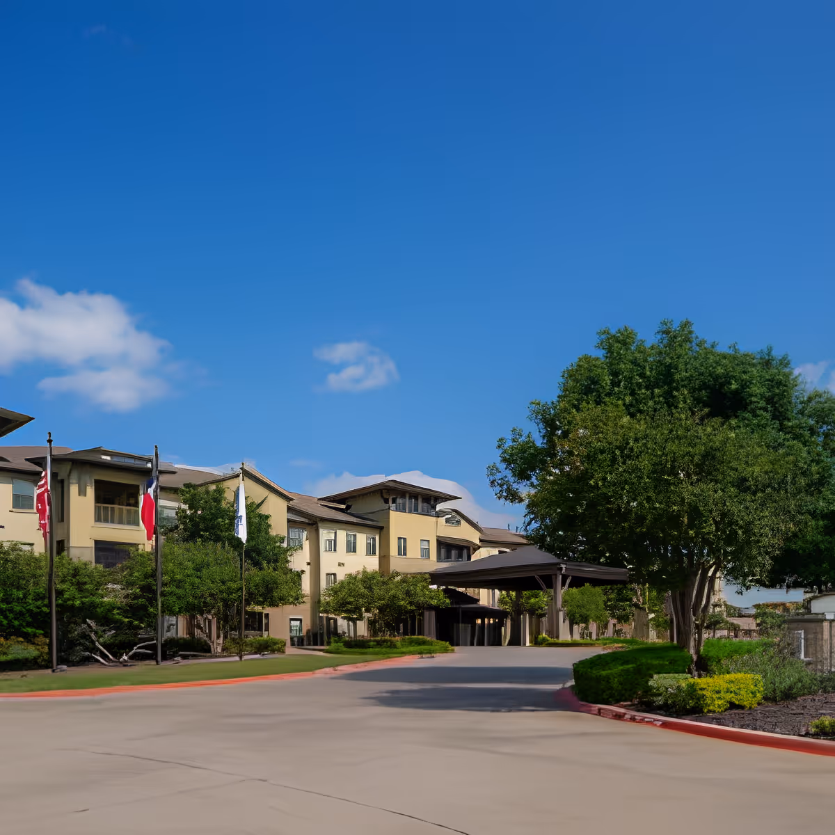Exterior view of Discovery Village At Twin Creeks, a multi-story senior living facility with a covered entrance, surrounded by trees and landscaping under a clear blue sky.