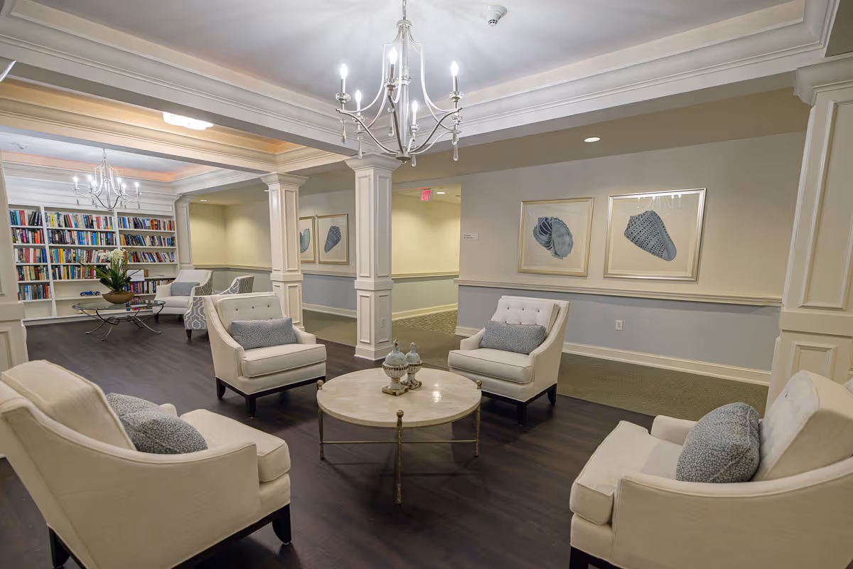 A cozy and elegant living room area in Marsh's Edge featuring four beige armchairs with gray cushions arranged around a round marble coffee table. The room has dark wood flooring, white pillars, and soft lighting from chandeliers. In the background, there is a bookshelf filled with books and two framed shell artworks on the wall.