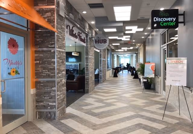 Interior hallway of a senior living facility with various rooms and centers along the corridor, including Maddy's Salon on the left, a Wellness Fitness room, The Hub, and a Discovery Center. The hallway has a patterned floor and ceiling lights, with chairs and tables visible further down the corridor.