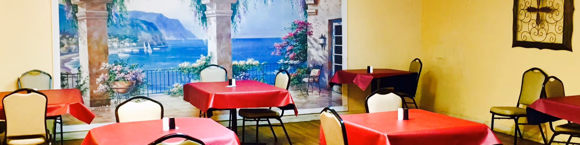 Dining area with several tables covered in red tablecloths and beige chairs. A large mural on the wall depicts a scenic coastal view with mountains, water, sailboats, and flowering plants.