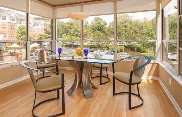Bright dining nook with a glass-top table and four chairs by large windows overlooking trees and a courtyard.