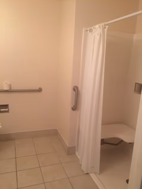 A bathroom with beige tiled floor and walls painted in light beige. There is a white shower area with a white curtain, a built-in bench, and a grab bar. A horizontal grab bar is mounted on the wall next to a toilet paper holder with a roll of toilet paper.