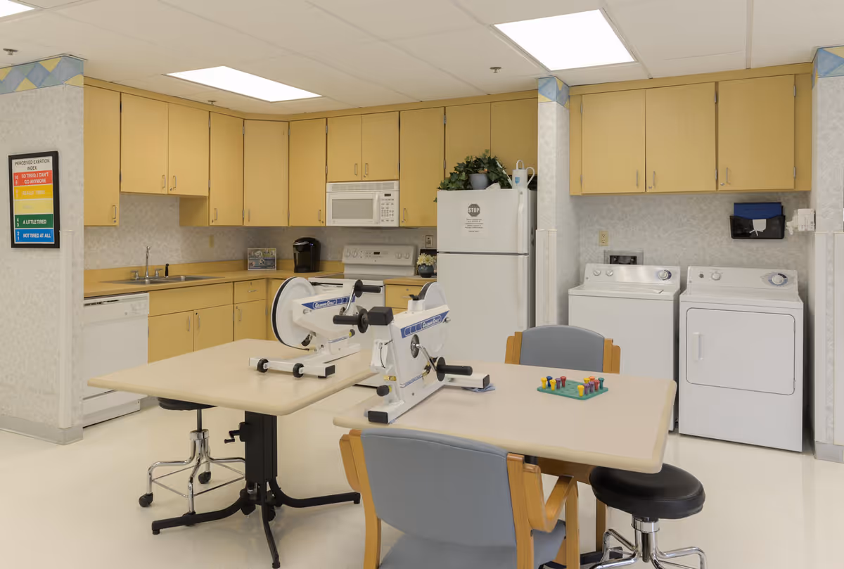 A multipurpose room in a senior living facility featuring a kitchenette with light wood cabinets, a microwave, dishwasher, and refrigerator. There is a table in the center with two hand cycle exercisers and a pegboard game. To the right, there is a washer and dryer set. The room has a clean, bright appearance with white walls and ceiling tiles.