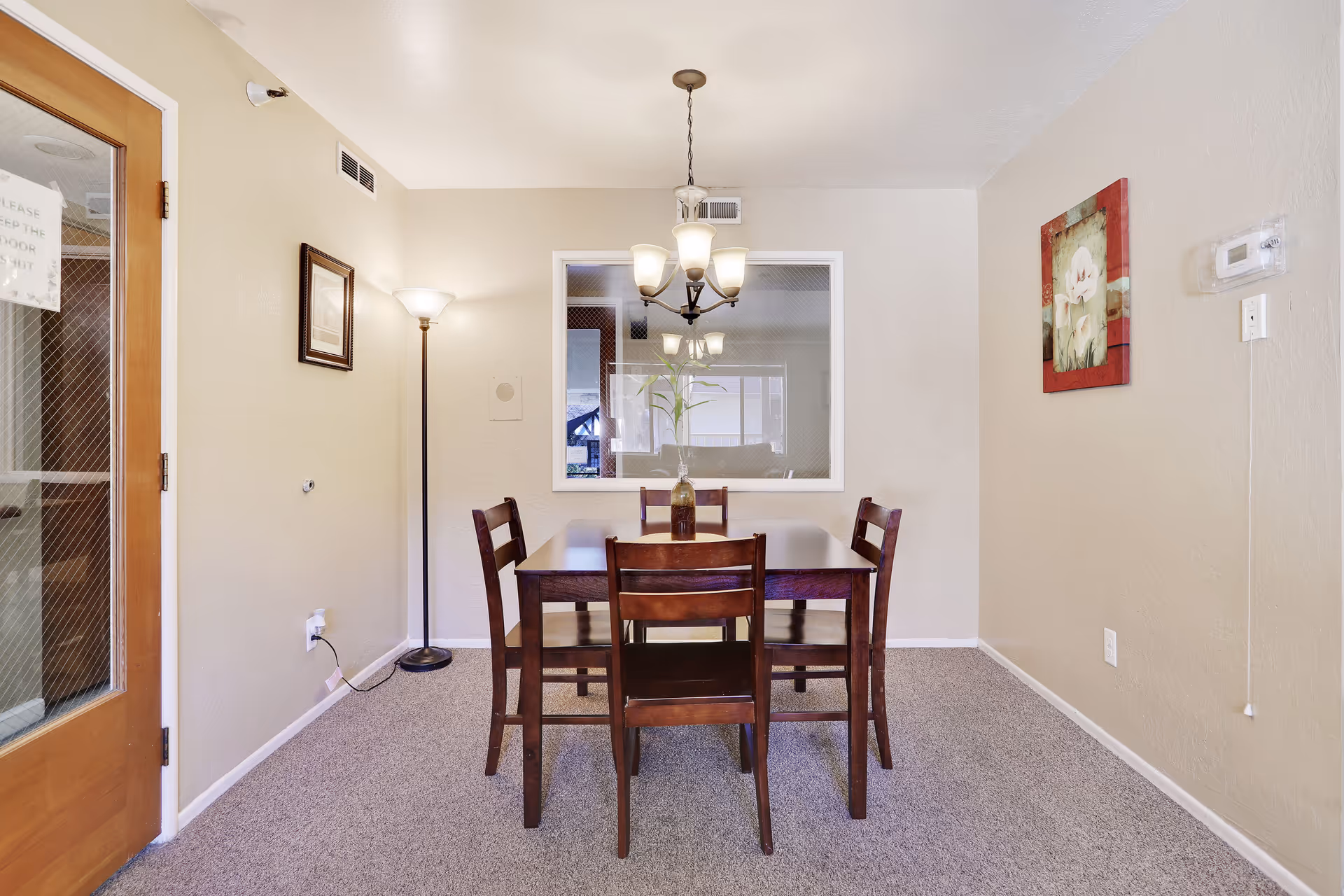 Wooden dining table with four chairs centered under a chandelier in a neutral-toned room with a floor lamp and wall art.