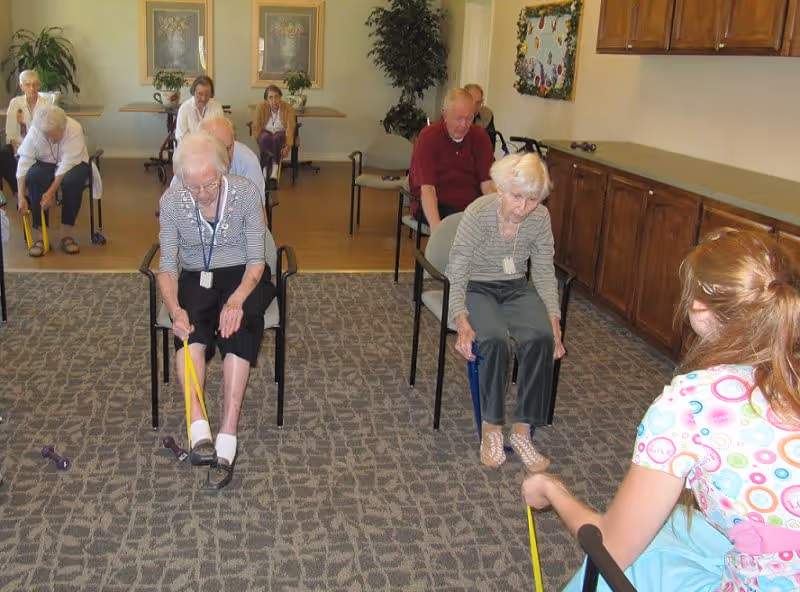 A group of elderly individuals seated in chairs in a room, participating in a seated exercise session using resistance bands. A caregiver or instructor is assisting them. The room has carpeted flooring, wooden cabinets along one wall, and framed artwork and plants in the background.