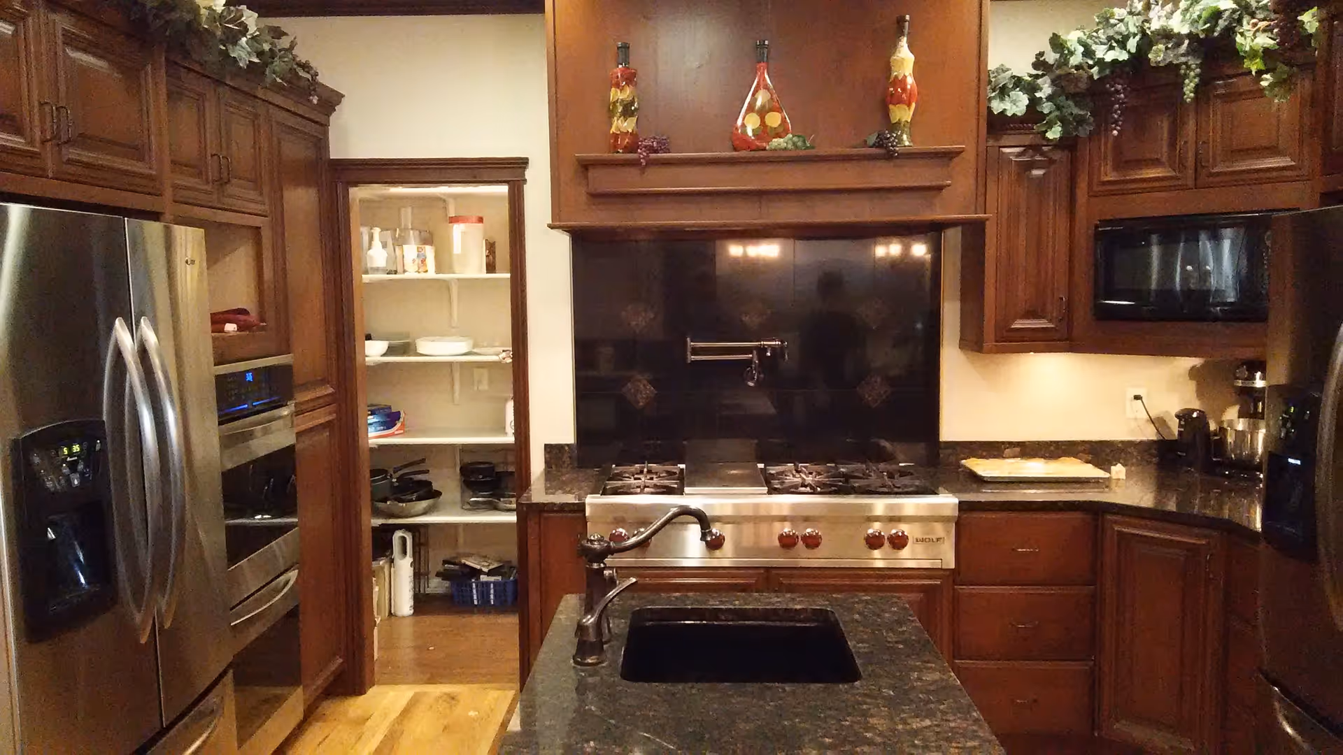 A kitchen with dark wooden cabinets, a stainless steel refrigerator, oven, and microwave. There is a black granite countertop with a sink and faucet in the foreground. A stove with red knobs and a black backsplash is centered in the image. The pantry door is open, showing shelves with various kitchen items. Decorative bottles and greenery are placed above the cabinets.