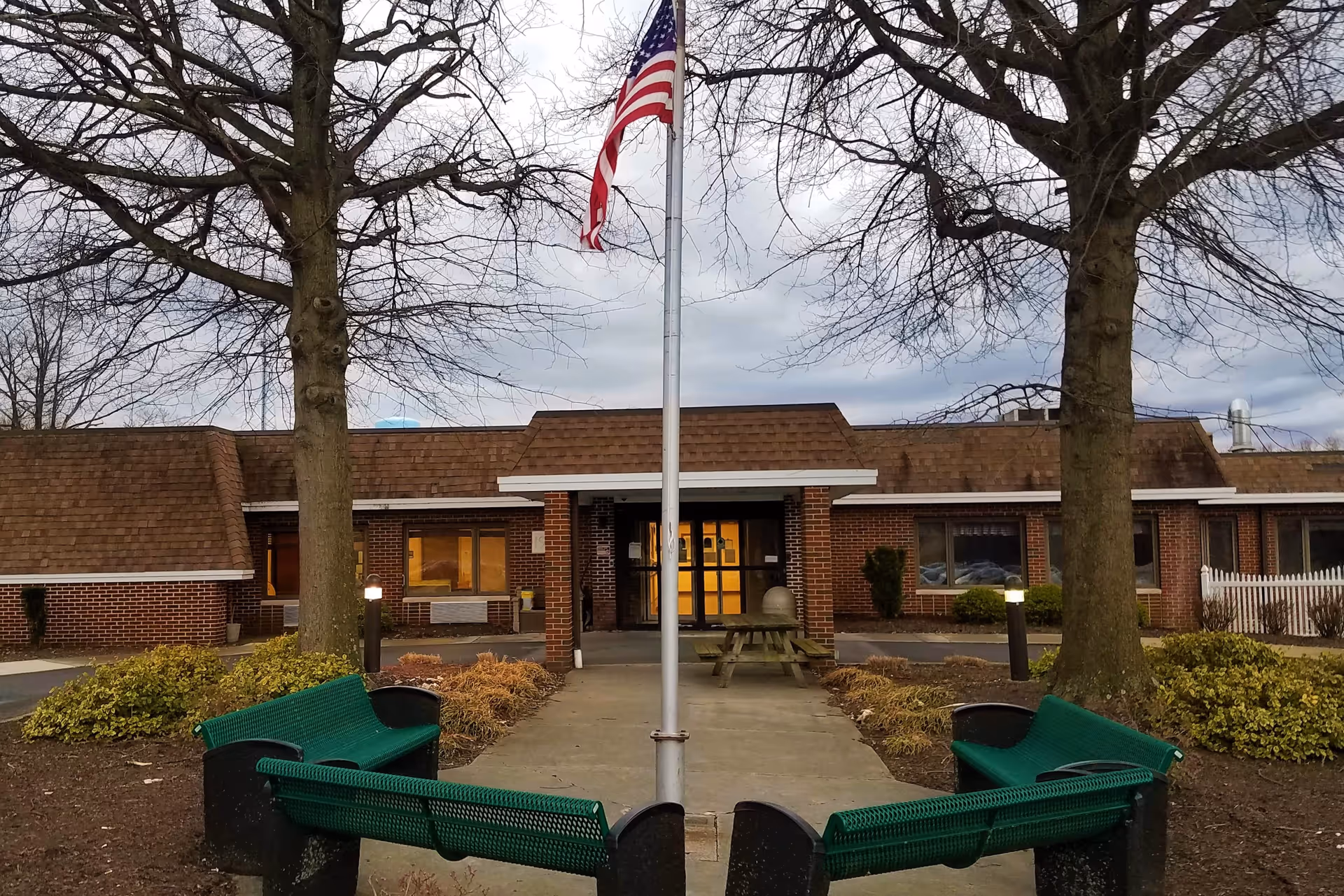 Front exterior view of Northern Dauphin Nursing & Rehabilitation building with a flagpole flying the American flag in the center. There are two large leafless trees on either side of the walkway leading to the entrance, green benches arranged around the flagpole, and some bushes and landscaping along the building.