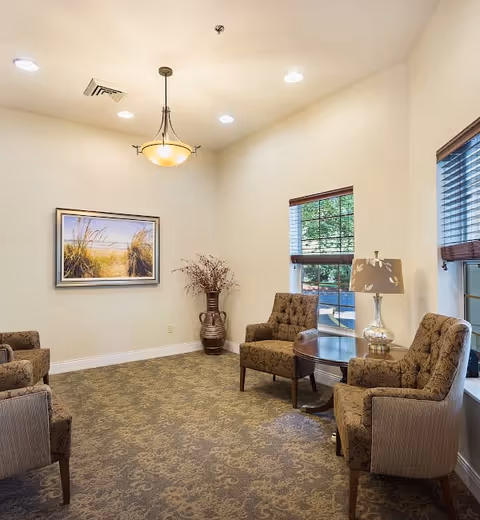 A cozy sitting area in Meadow Creek Village Assisted Living featuring four upholstered armchairs arranged around a round wooden table with a decorative lamp. The room has beige walls, a patterned carpet, a large window with blinds, a framed landscape painting, and a tall vase with dried branches in the corner.