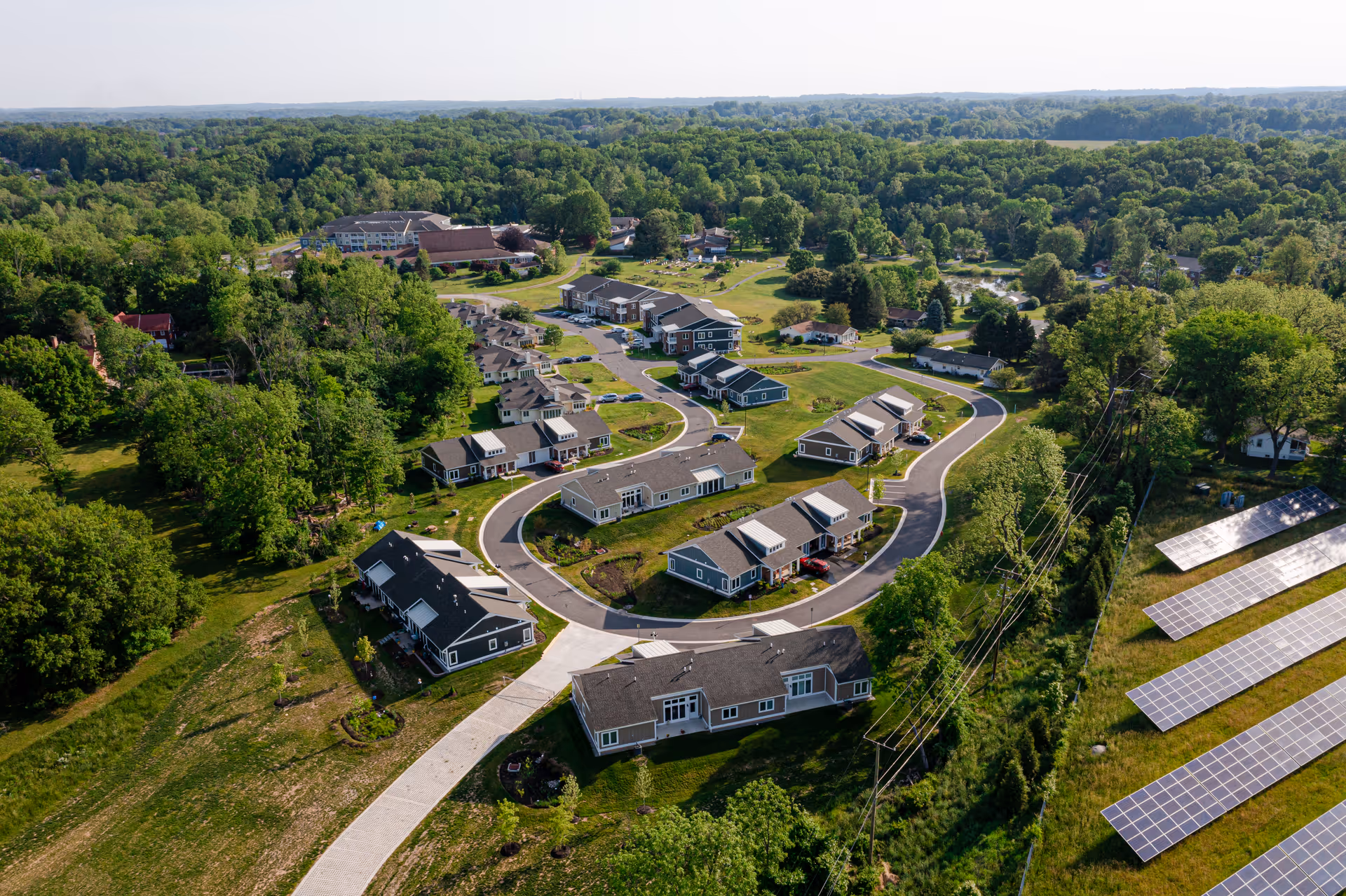 Aerial view of Friends House Retirement Community showing multiple single-story residential buildings arranged along curved roads surrounded by green lawns and trees, with solar panels visible on the right side.