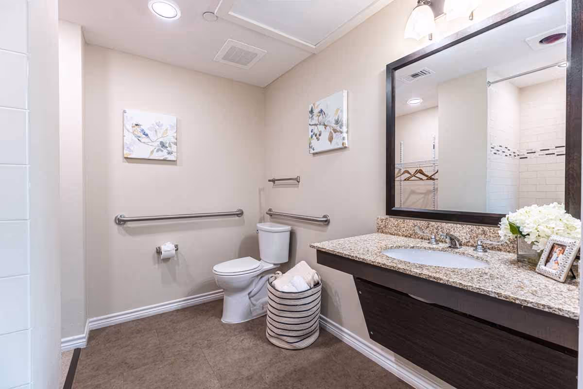 A clean and modern bathroom with beige walls and a tiled floor. There is a white toilet with grab bars on both sides, a granite countertop with a sink, a large mirror above the sink, and a basket with rolled towels on the floor. A framed picture and a vase with white flowers are on the countertop. A shower area with white tiles is partially visible on the left.