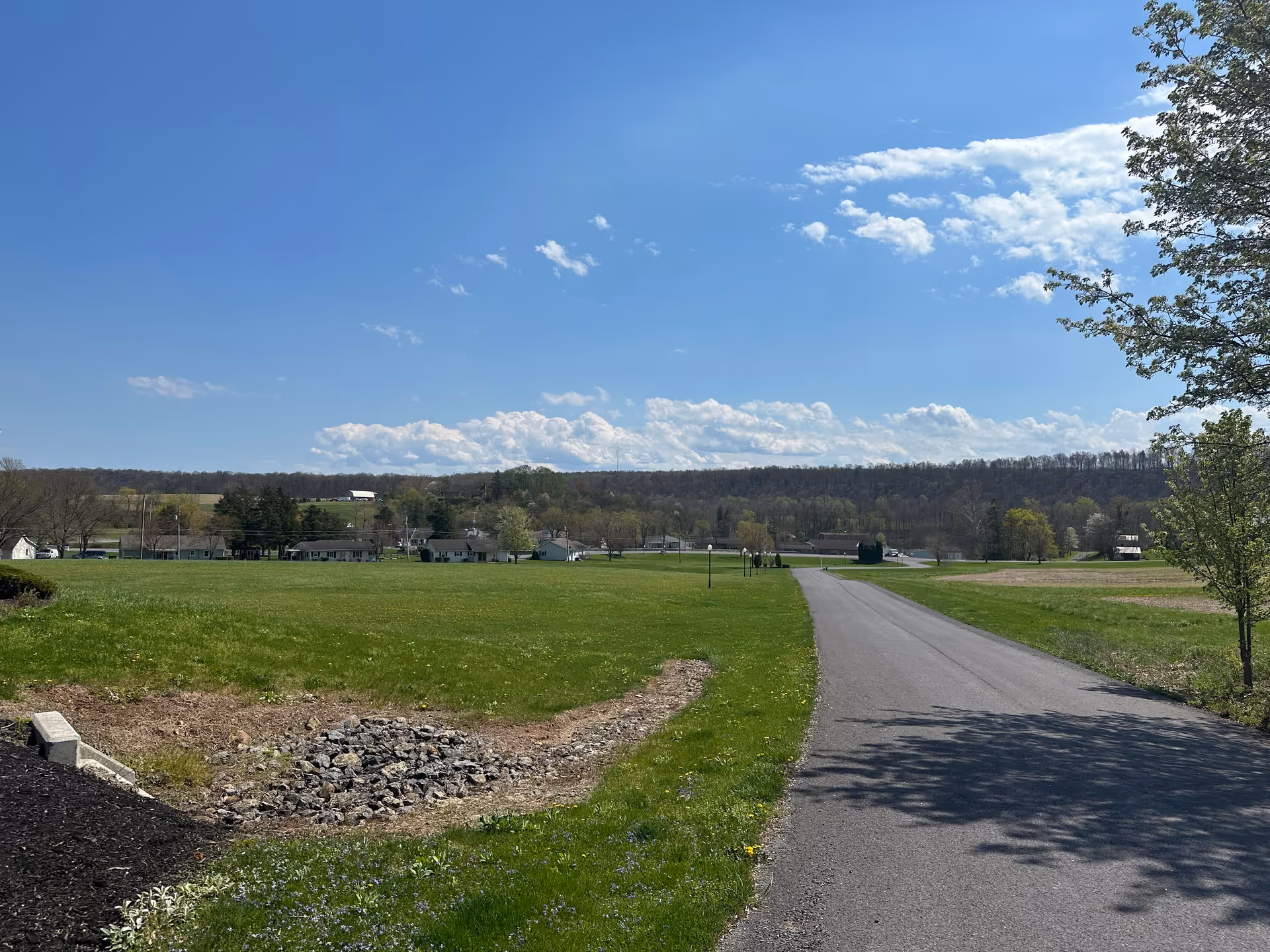 A paved road runs through a large grassy field with some trees on the right side. In the distance, there are houses and a wooded hill under a blue sky with scattered clouds.