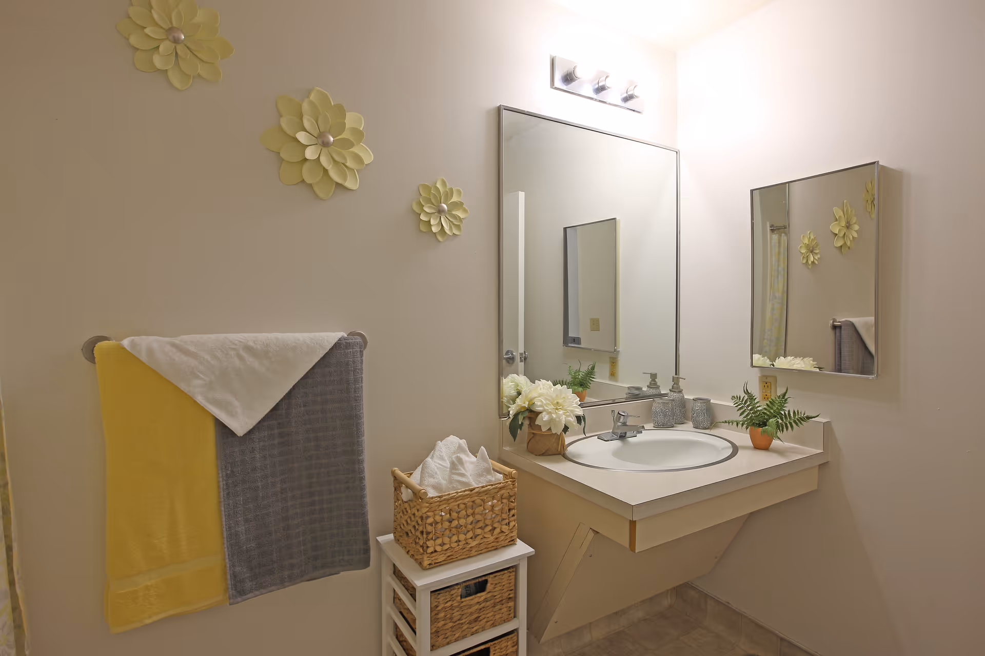 A clean bathroom with a wall-mounted sink and a large mirror above it. The sink countertop has decorative items including a small potted plant and a basket with tissues. On the wall to the left, there are three yellow flower-shaped wall decorations and a towel rack holding a yellow and a gray towel. The floor has a light-colored tile, and the walls are painted a soft beige.