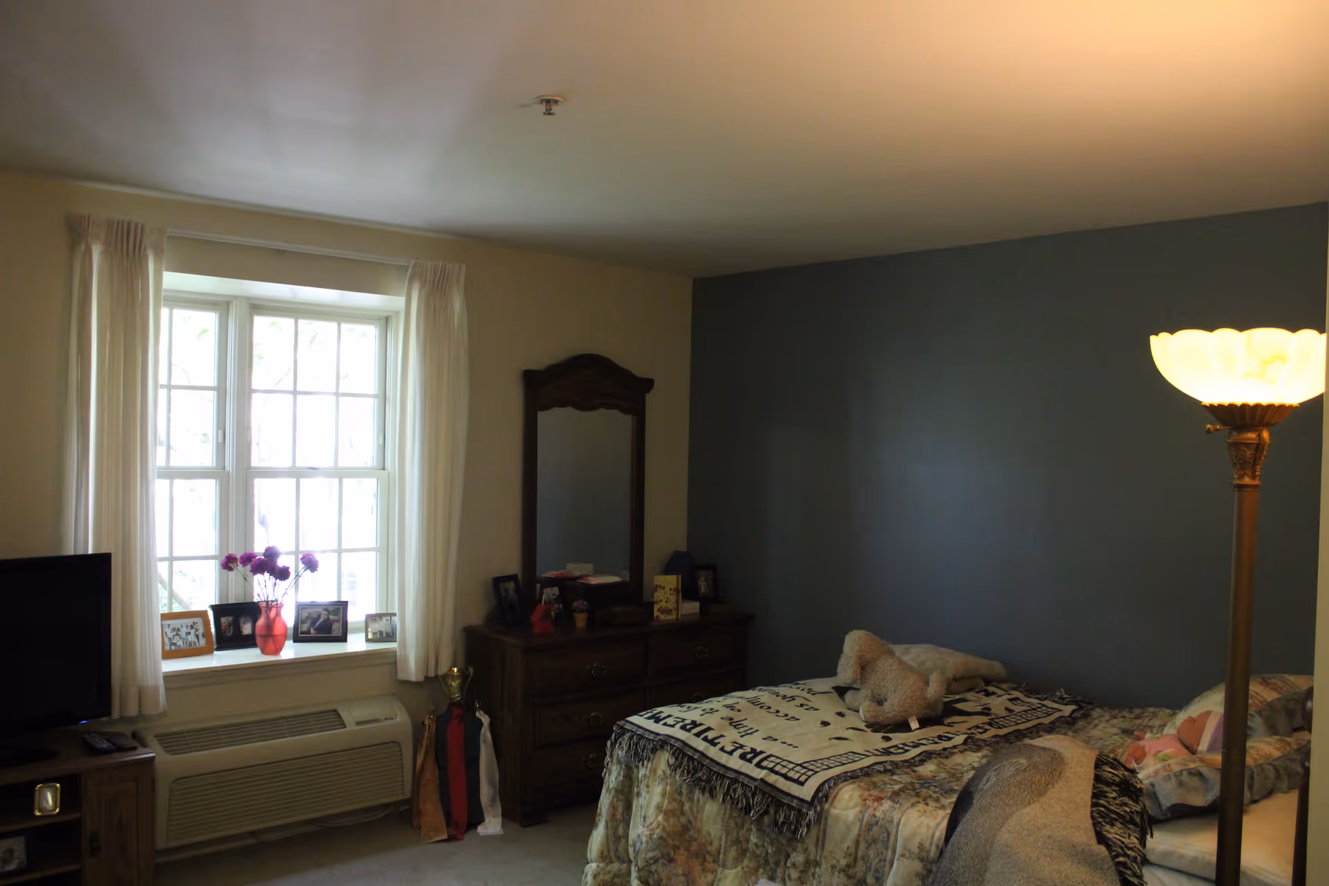 A cozy bedroom with a bed covered in patterned blankets and a stuffed animal on top. There is a wooden dresser with a mirror against a blue accent wall. A window with white curtains lets in natural light, and a floor lamp with a glowing shade stands near the bed. Several framed photos and a vase with flowers are on the windowsill and dresser.