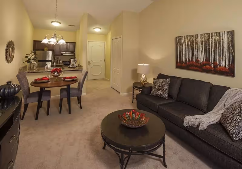Open-concept living room and dining area with a dark sofa, round coffee table, small dining table, and a kitchen visible in the background.