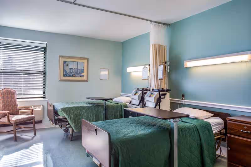 A hospital-style room with two beds covered in green blankets, each equipped with an overbed table and medical equipment mounted on the wall. There is a wooden chair near a window with blinds, a framed picture on the wall, and a small wooden nightstand beside one of the beds. The walls are painted light blue and the room is well-lit with natural and artificial light.