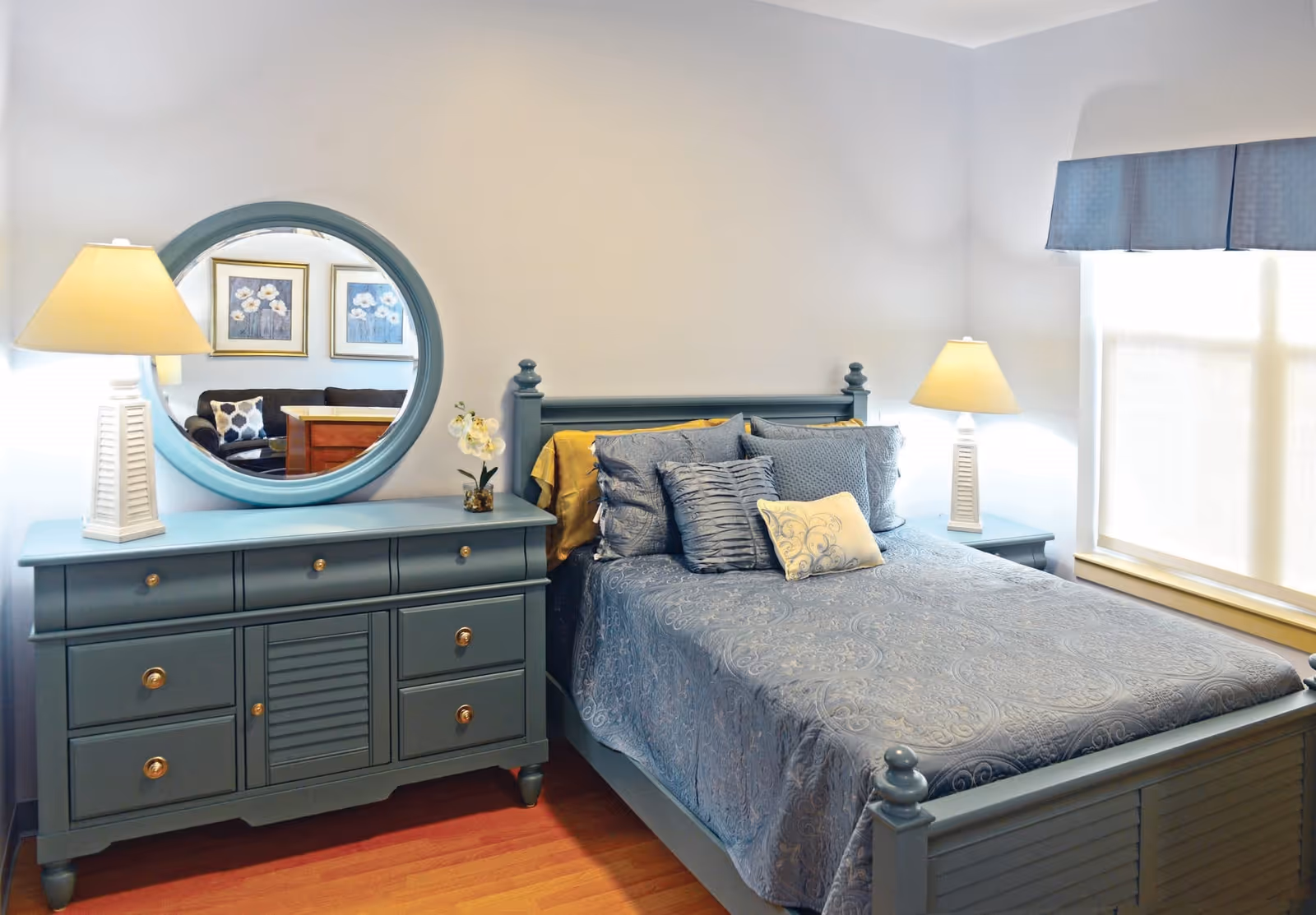 Well-lit bedroom with a blue wooden bed, matching dresser with round mirror, and bedside lamps.