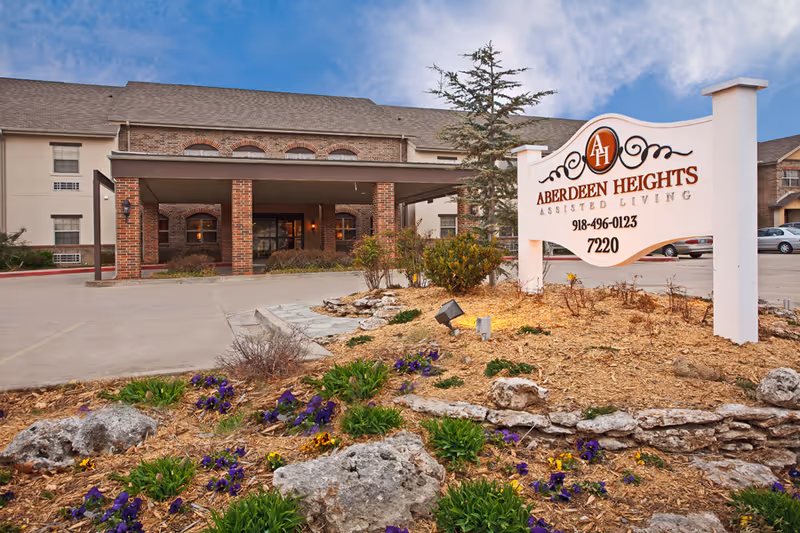 Exterior view of Aberdeen Heights Assisted Living facility showing the main entrance with a covered driveway, a landscaped garden with flowers and rocks in the foreground, and a large white sign displaying the facility name, phone number, and address.