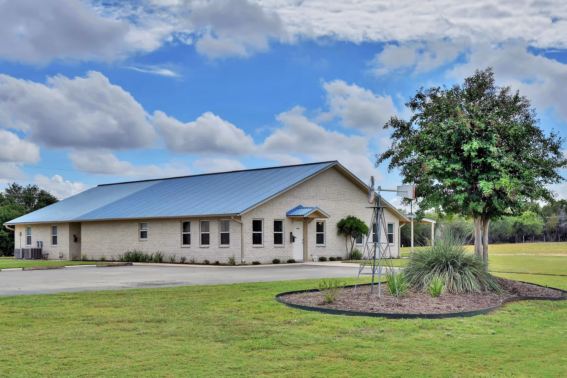 A single-story building with a light-colored brick exterior and a blue metal roof, surrounded by a well-maintained lawn and landscaping including a tree and ornamental grasses. The sky is partly cloudy with patches of blue.