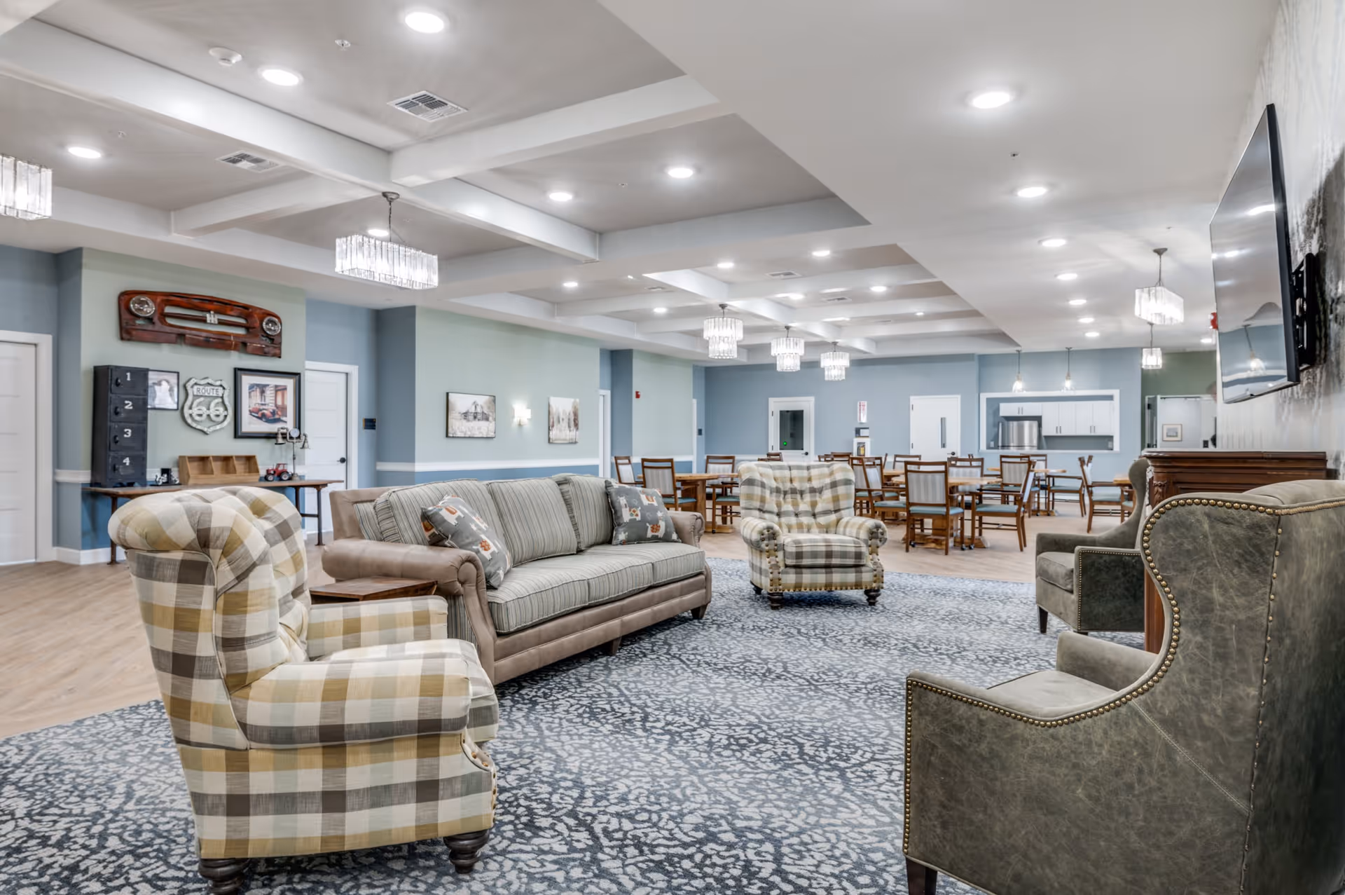 A spacious and well-lit common area in a senior living facility featuring comfortable seating including plaid and leather armchairs and a sofa arranged on a patterned carpet. In the background, there are multiple wooden dining tables and chairs under modern chandeliers, with a kitchen area visible further back. The walls are painted in soft blue and green tones, decorated with framed pictures and vintage-style decor.