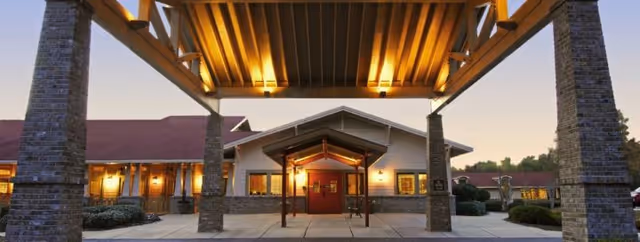 Entrance of a senior living facility with a covered drop-off area supported by stone pillars, warm lighting under the roof, and a building with a peaked roof and double doors in the background during dusk.