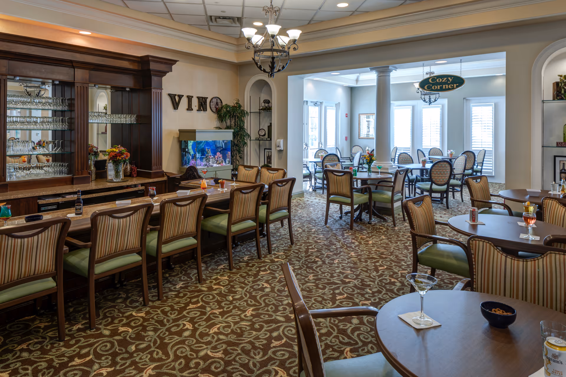 Interior view of a senior living facility's dining and lounge area featuring a long bar with high chairs, tables with chairs arranged for dining, a fish tank, and decorative elements including a sign that reads 'VINO' and another sign that says 'Cozy Corner'. The room has patterned carpet, chandeliers, and large windows with blinds allowing natural light.