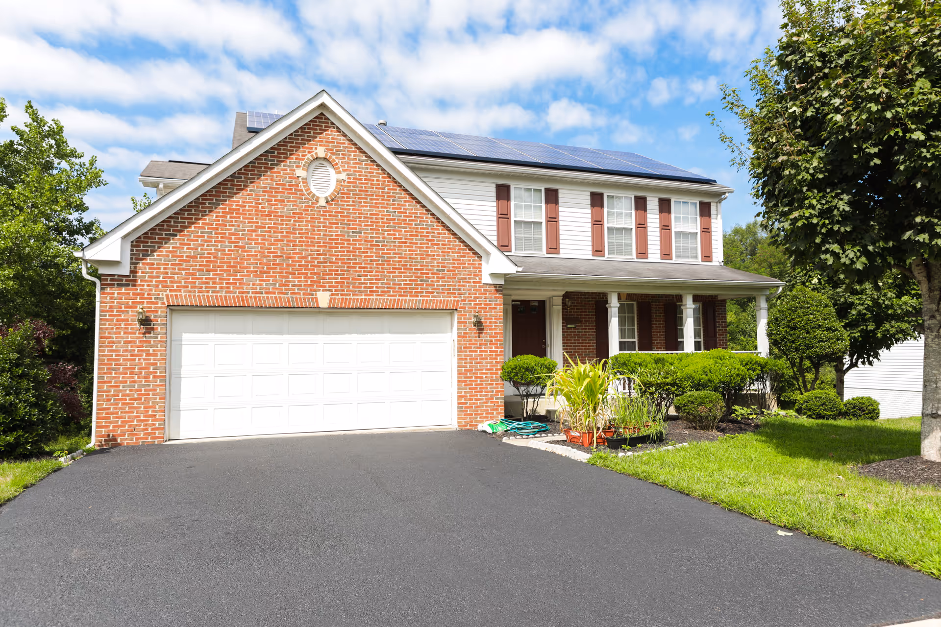Front exterior view of a two-story house with a brick facade and white garage door. The house has solar panels on the roof, red shutters on the windows, a covered porch with white columns, and a well-maintained garden with bushes and trees. The driveway is freshly paved and the sky is partly cloudy.