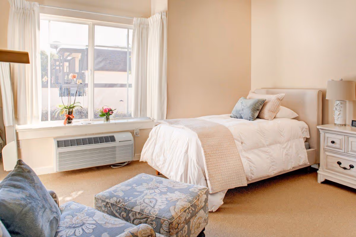 Sunny, softly decorated single bedroom with a made twin bed, nightstand, window seat and upholstered chair.