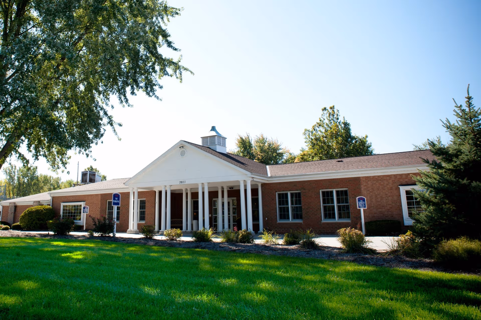 Exterior view of Glenbrook Rehabilitation and Skilled Nursing Center, a single-story brick building with white columns at the entrance, surrounded by green grass, trees, and shrubs under a clear blue sky.