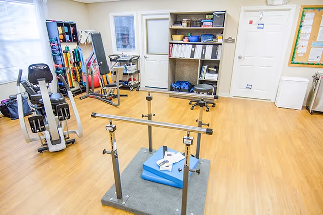 A bright room with wooden flooring equipped for physical therapy or exercise, featuring parallel bars in the center, exercise machines, weights, and shelves with various supplies and equipment. There are two doors and a bulletin board on the walls.