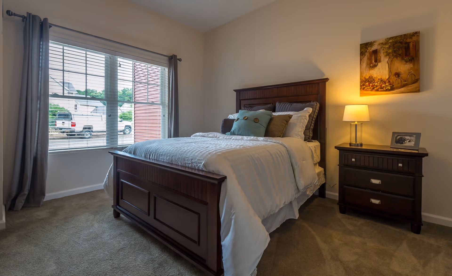 A cozy bedroom with a large wooden bed frame and a neatly made bed with white and gray bedding and decorative pillows. Next to the bed is a dark wooden nightstand with a lamp and a framed photo. A painting hangs on the wall above the nightstand. A large window with gray curtains lets in natural light and shows a view of parked vehicles outside.
