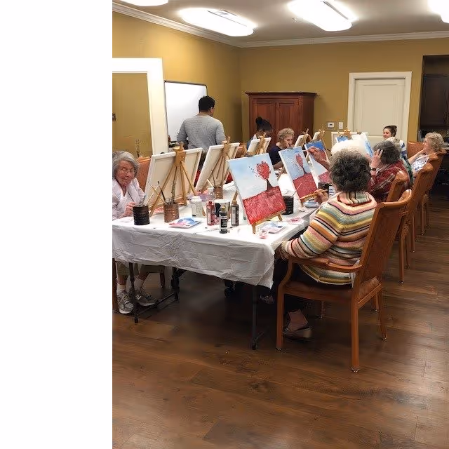 A group of elderly individuals seated around a long table in a room, engaged in a painting activity with canvases on easels, paint bottles, and brushes. The room has wooden flooring, yellow walls, and overhead fluorescent lighting.