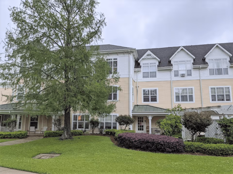 Front exterior of a three-story light-yellow residential building with a covered entrance, large tree, and manicured lawn.