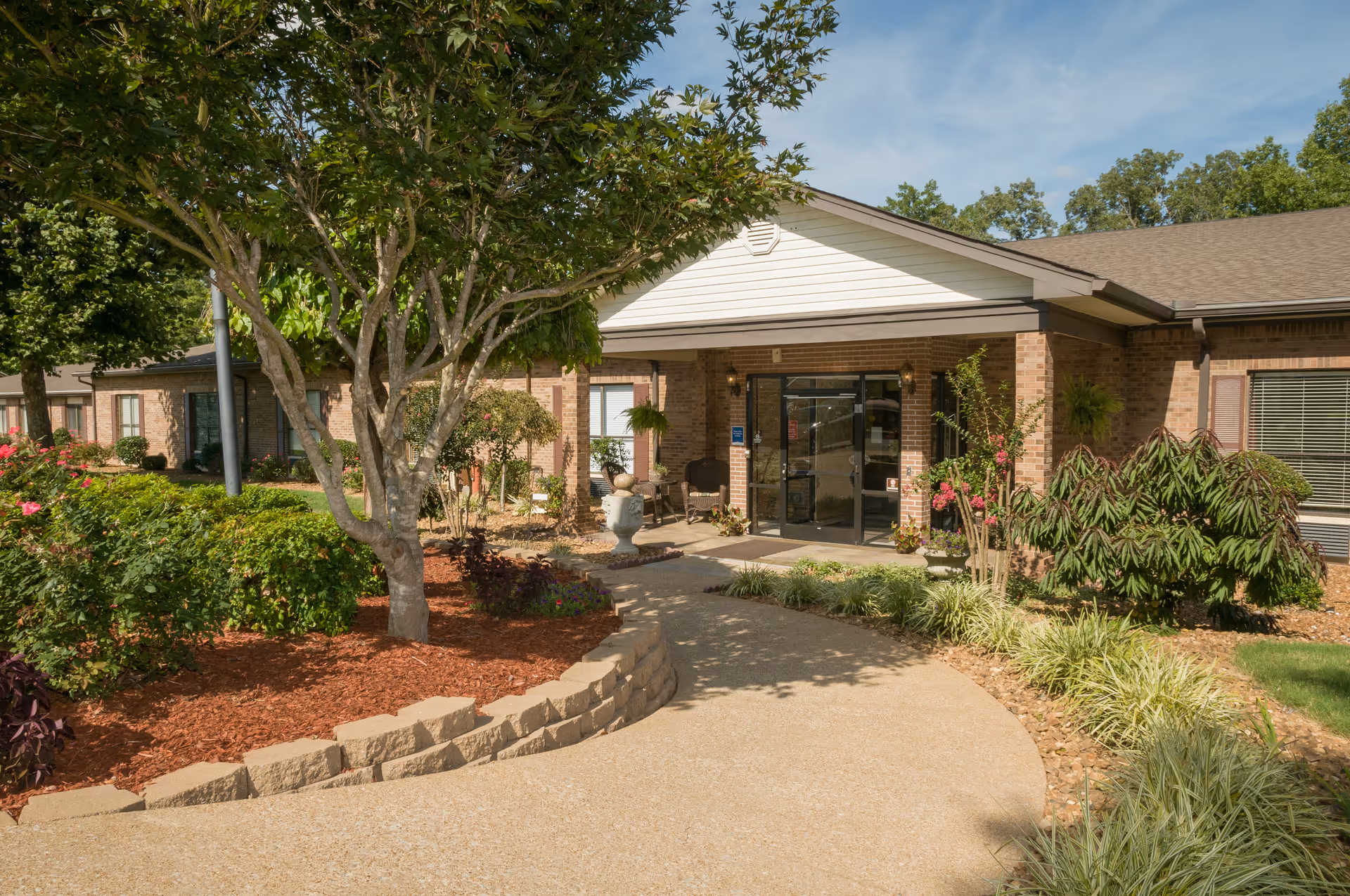Front entrance of a single-story brick senior living facility with a curved walkway and landscaped gardens.
