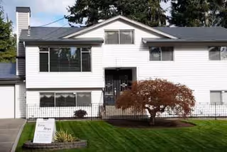 Front view of a two-story white residential building with a manicured lawn, small tree, and a sign in the yard.