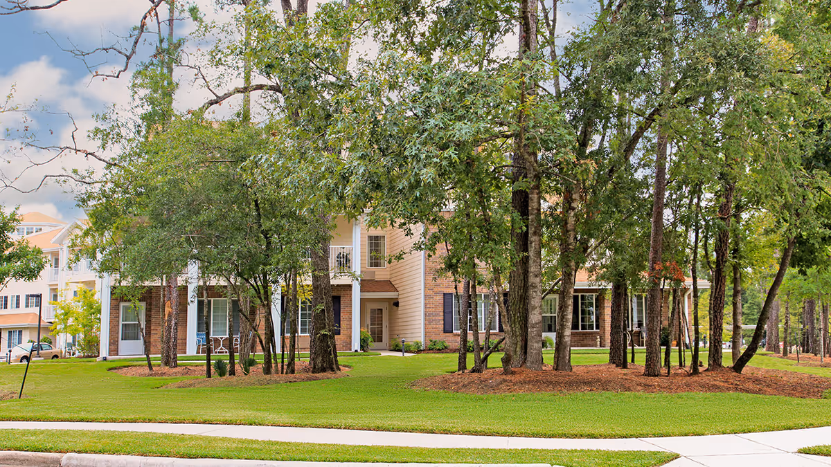 Exterior view of a senior living facility building partially obscured by tall trees and greenery, with a well-maintained lawn and sidewalk in the foreground under a partly cloudy sky.