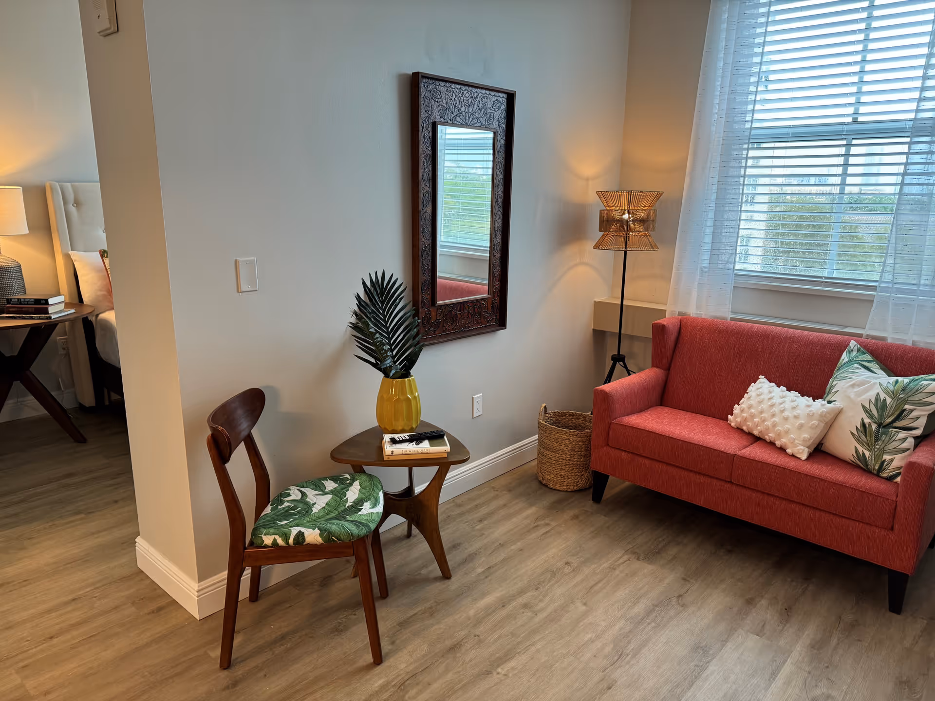 A cozy living area with a red loveseat adorned with two decorative pillows, one white with textured dots and the other with a green leaf pattern. Next to the loveseat is a tall floor lamp with a woven lampshade and a wicker basket. A wooden chair with a green leaf-patterned cushion sits beside a small wooden side table holding a yellow vase with green leaves and two books. A decorative mirror with a carved wooden frame hangs on the wall above the table. The room has light wood flooring and a large window with white blinds and sheer curtains, letting in natural light. A partial view of a bedroom with a bed and nightstand with a lamp is visible through an open doorway.