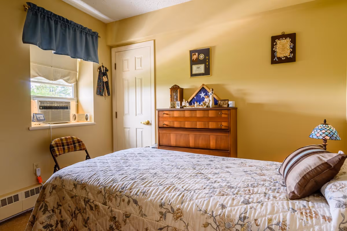 A cozy bedroom with a bed covered in a floral quilt and a striped pillow. There is a wooden dresser against the wall with framed photos, a folded American flag in a display case, and decorative items on top. A window with an air conditioning unit and a blue valance is on the left side, with a plaid chair beneath it. The walls are painted light yellow, and there is a small Tiffany-style lamp on a bedside table.