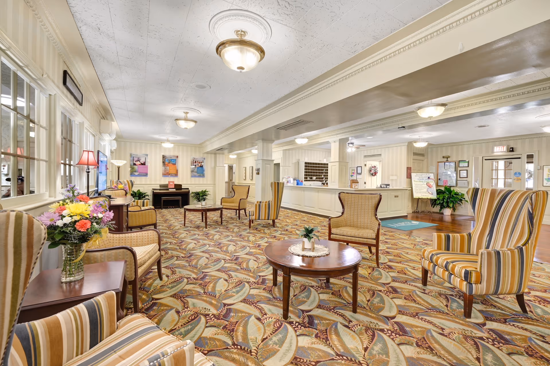 A spacious and well-lit senior living facility lobby with patterned carpet, multiple comfortable armchairs with striped upholstery, round wooden tables with small plants and flowers, a reception desk in the background, and framed artwork on the walls.