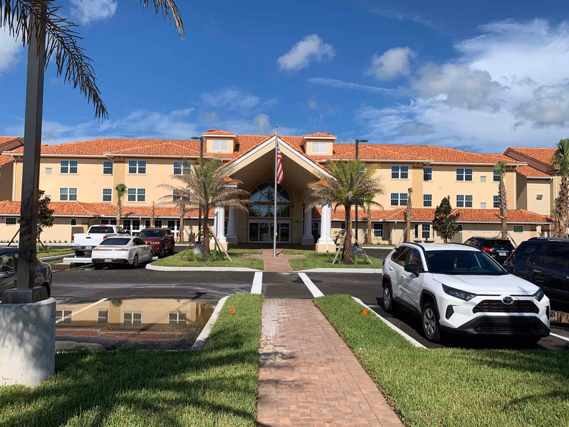 Front exterior view of Shell Harbor Retirement Community building with a red-tiled roof, beige walls, palm trees, an American flag on a flagpole, a paved walkway leading to the entrance, and several parked cars in the parking lot under a partly cloudy blue sky.