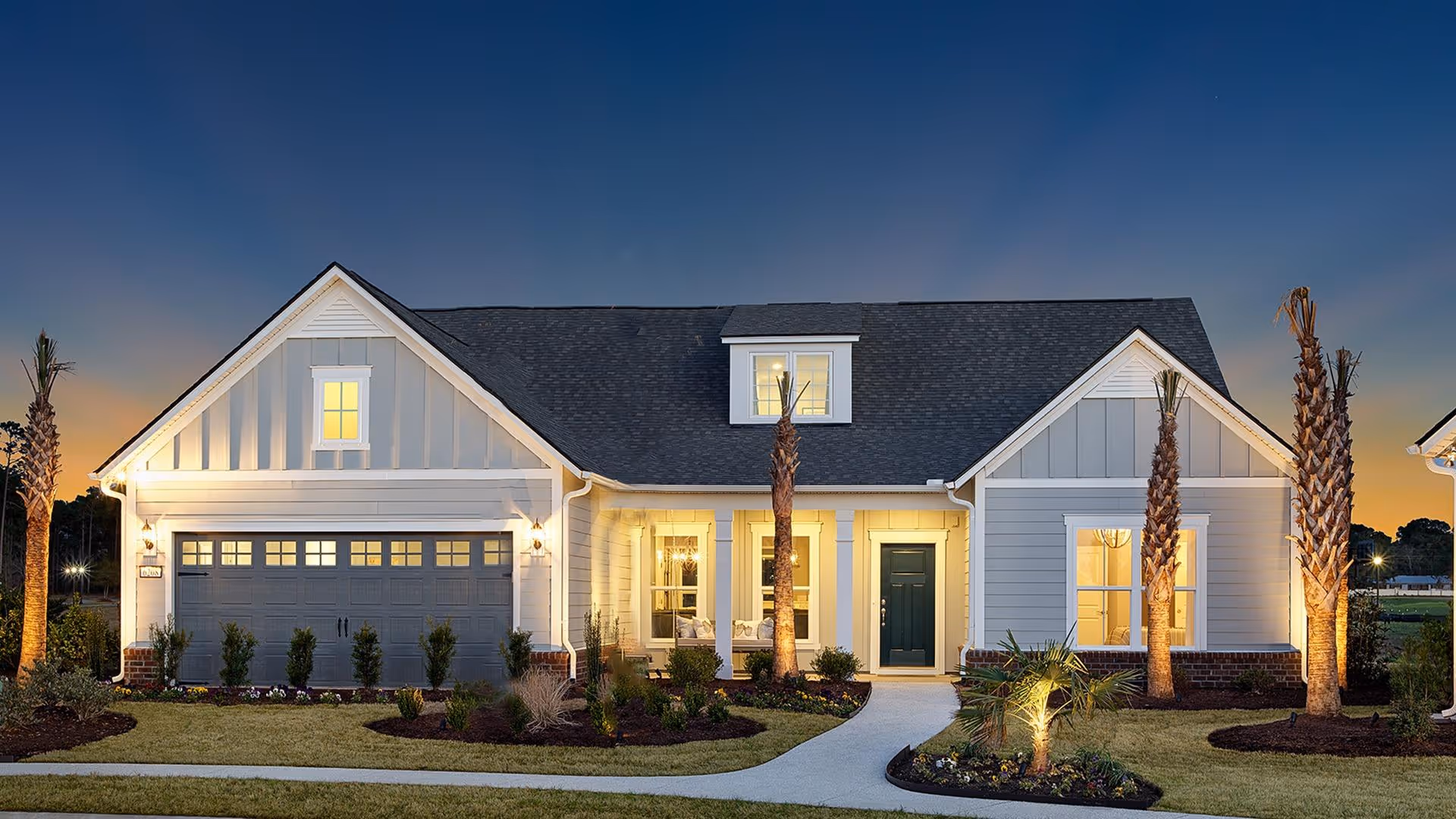 Exterior view of a single-story house at dusk with a well-lit front porch, a two-car garage, and landscaped yard featuring palm trees and shrubs.