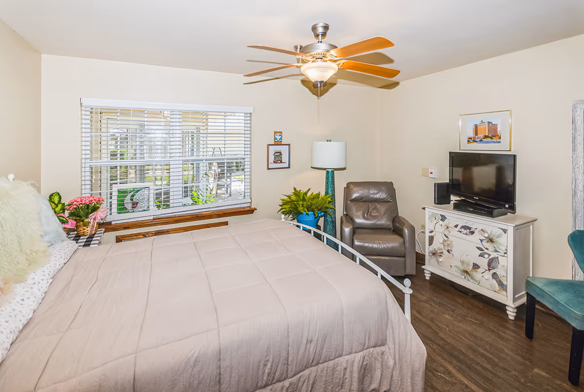 A cozy bedroom with a large bed covered in a beige comforter, a window with white blinds, a ceiling fan with wooden blades, a brown leather armchair, a floral painted dresser with a TV on top, a green chair, and several potted plants and framed pictures on the walls.