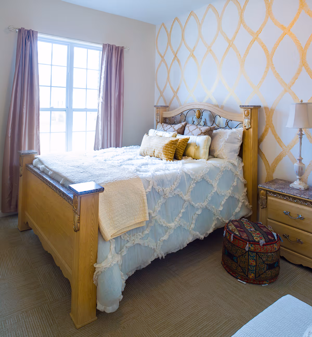 A cozy bedroom with a wooden bed frame featuring intricate metalwork on the headboard. The bed is made with a light blue and white ruffled bedspread and several decorative pillows in shades of gold and beige. There is a window with pink curtains letting in natural light. Next to the bed is a wooden nightstand with a marble top and a lamp. A colorful patterned ottoman is placed on the carpeted floor beside the nightstand. The wall behind the bed has a gold geometric pattern.